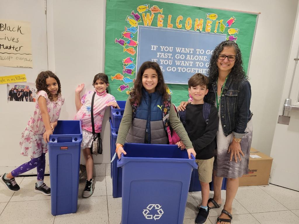 A group of children and their teacher smile with a recycling bin