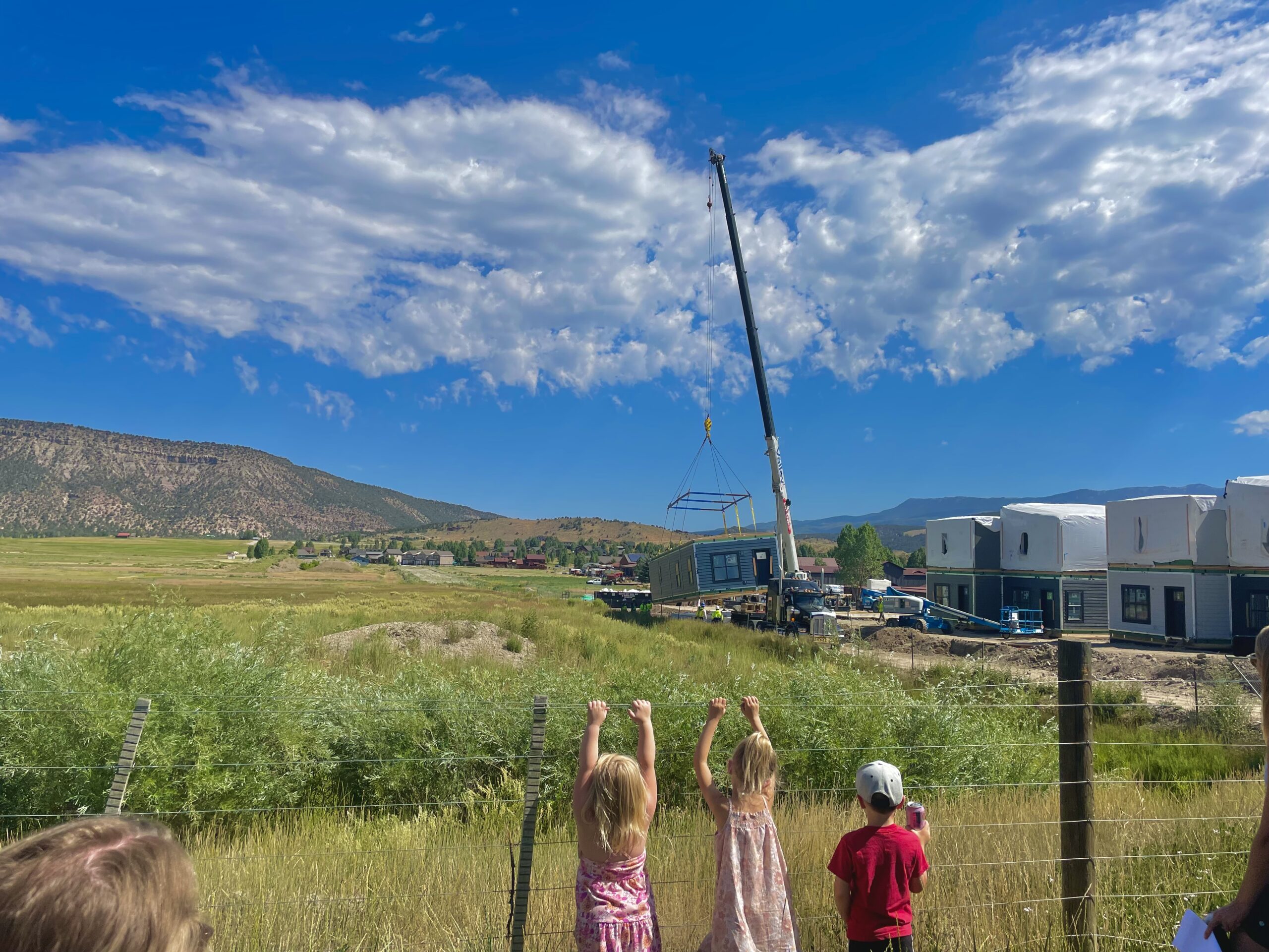 A group of children stand against a fence watching a home being built