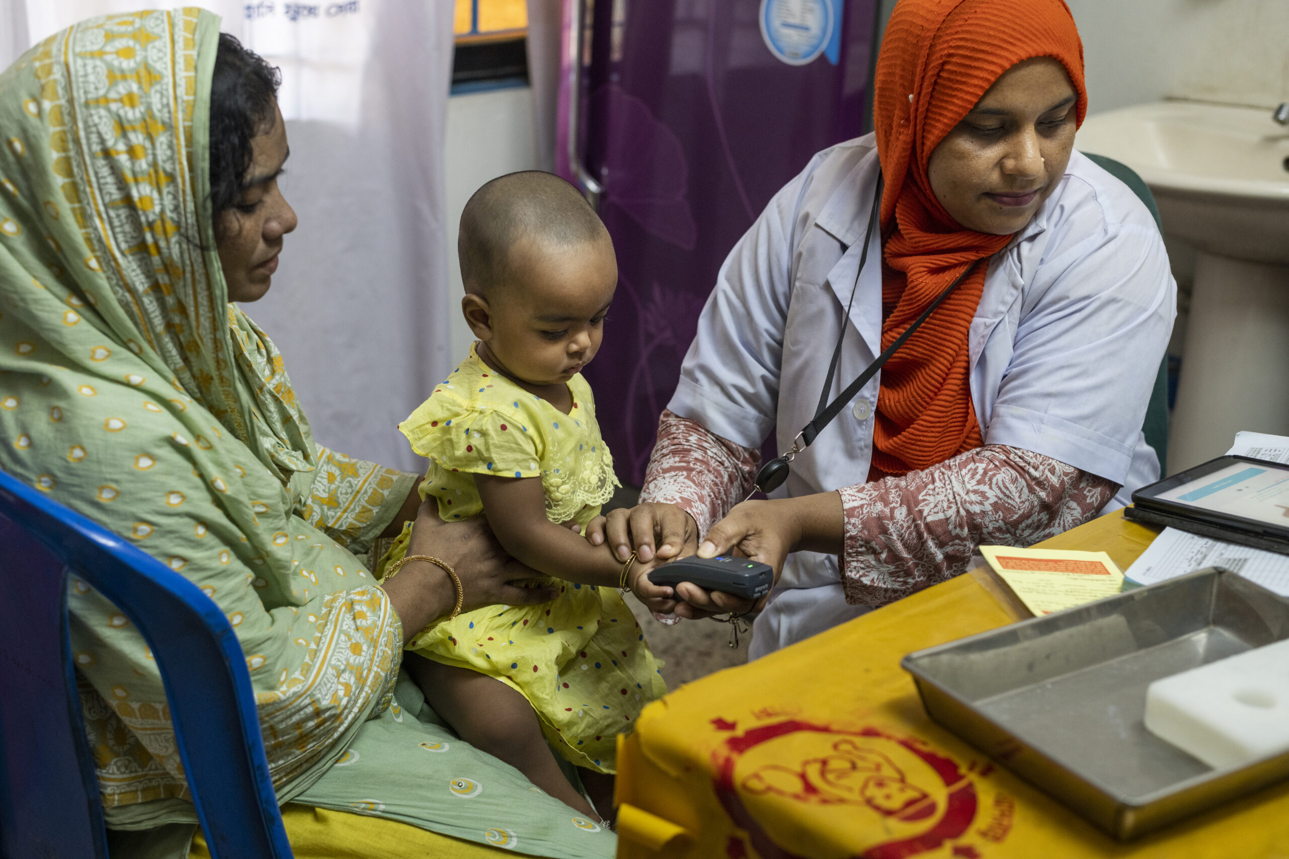 An aid worker presses a child's finger to a fingerprint scanner while the child sits on their mother's lap.